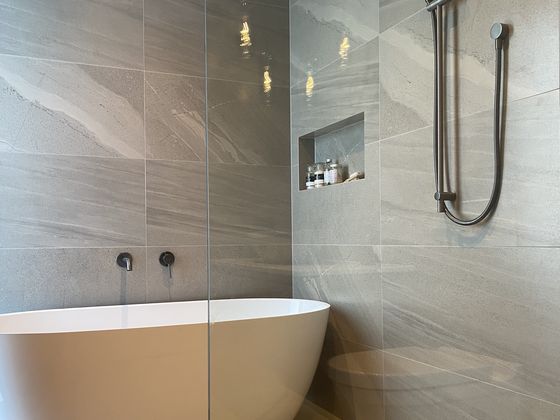 A freestanding bath beside a glazed walk-in shower, with black wall-mounted tapware, a hand shower on a rail and a tiled recess set into the wall.