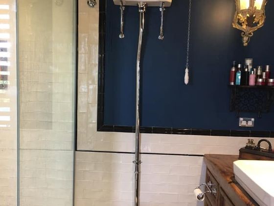 A high-level cistern toilet with a black seat and a chrome flush pipe against a deep navy wall, beside a glass shower screen.