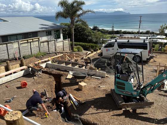 Two GD Plumbing Services plumbers working in the foundation trenches of a coastal new build, with a digger and a branded van on the site behind them.