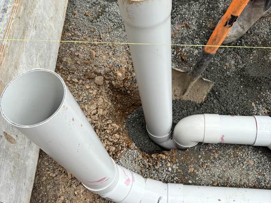 Two white PVC drainage risers and a swept junction bend set in the gravel of a new build foundation, ready for the slab to be poured over them.