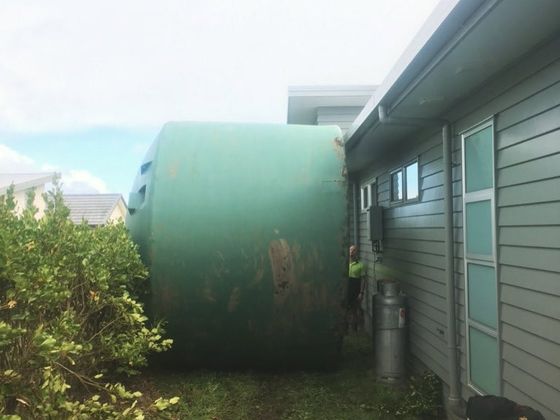 A large green water tank being worked down the grassed side of a house, past the guttering and downpipes.