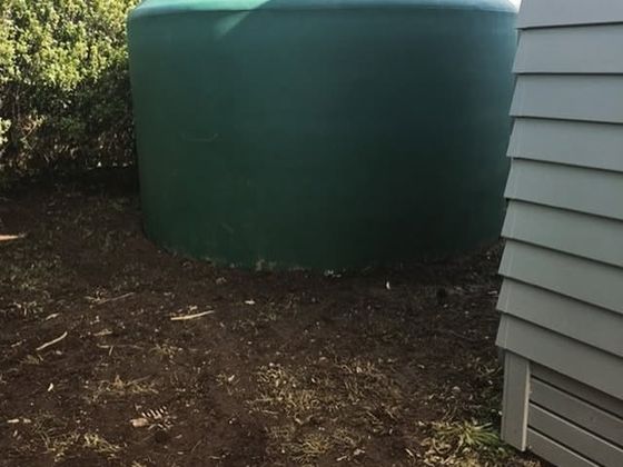 A new green water tank standing on freshly turned soil beside a weatherboard house.