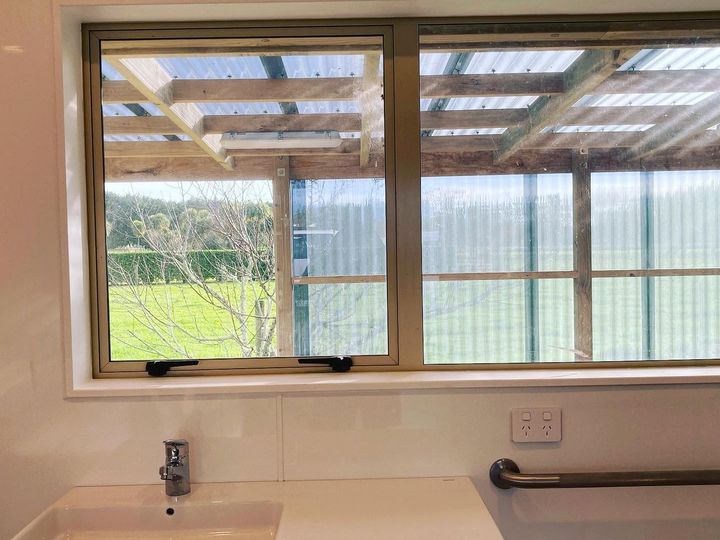 A wall-hung basin with a lever mixer and a stainless steel grab rail, under a timber-framed window looking out to paddocks.